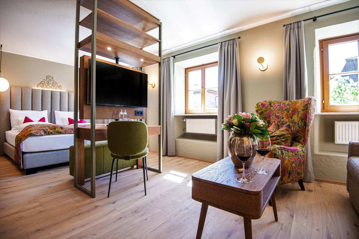 Habitación de hotel moderna con cama, televisor, una silla verde, una mesa con copas de vino y una ventana con cortinas.