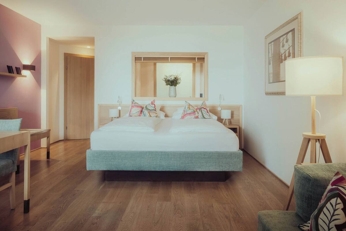 Habitación de hotel con cama, mesitas de noche, escritorio, silla, lámpara y ventana con planta.