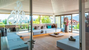 A modern interior with a wooden ceiling, a seating area with cushions, a large window, and a view of greenery and buildings.