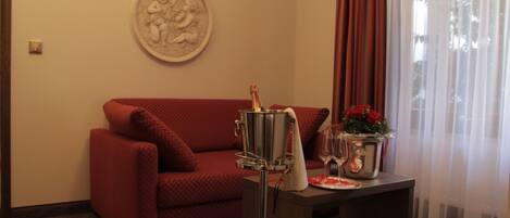 Una habitación con un sofá rojo, una pequeña mesa con una botella de champán, un florero con flores rojas y un plato decorativo colgado en la pared.