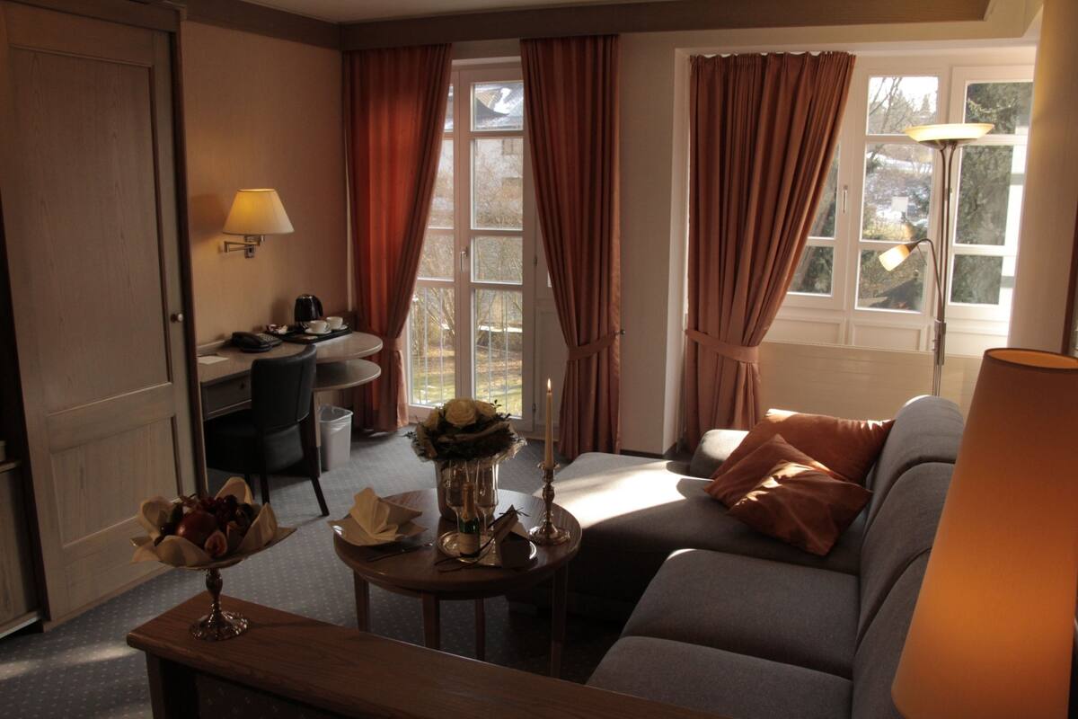Una sala de estar con un sofá, una mesa con un florero y velas, un escritorio con una lámpara, y vistas a través de una ventana con cortinas.