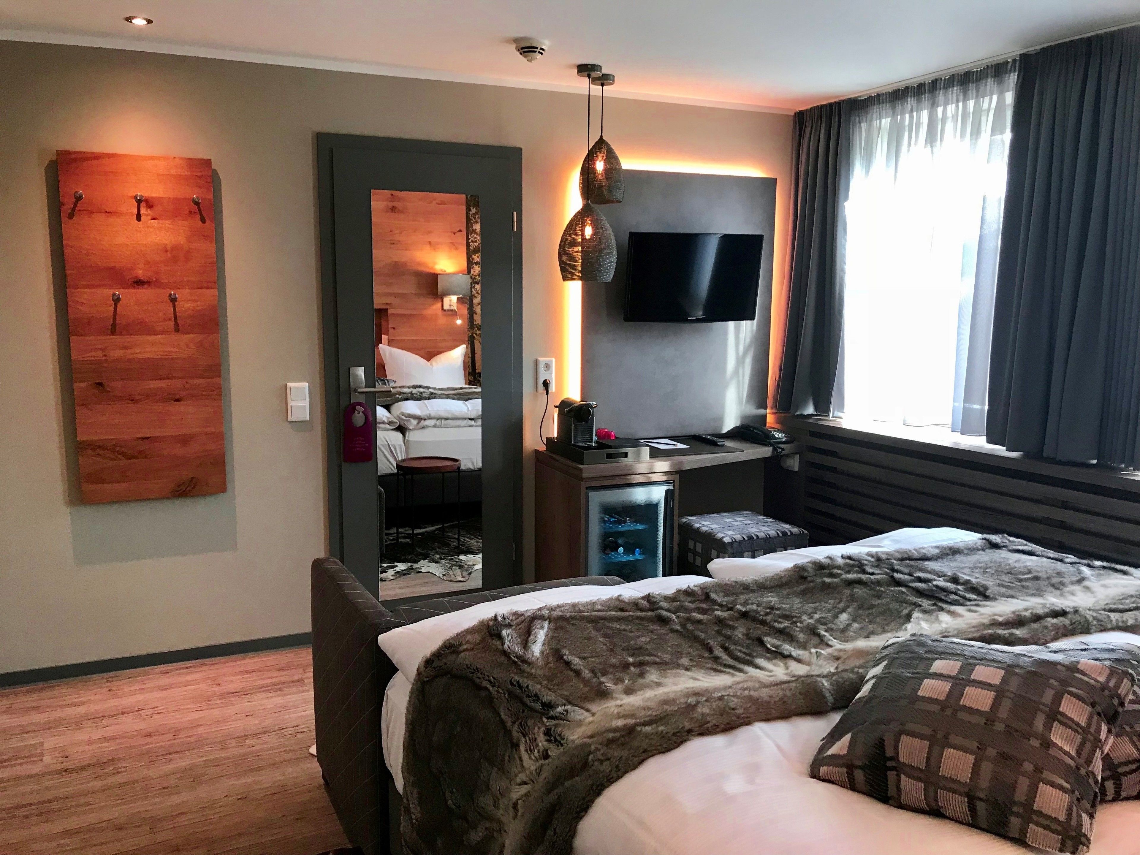 Ein modernes Hotelzimmer mit einem großen Bett, einem flachbildfernseher an der Wand, einem kleinen Schreibtisch mit Stuhl und einem großen Fenster mit Vorhängen.