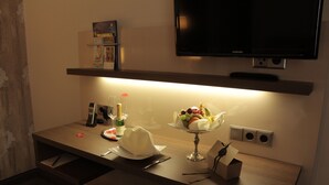 Una habitación de hotel moderna con un escritorio de madera, un televisor de pantalla plana montado encima y una pequeña estantería con libros y un objeto decorativo.