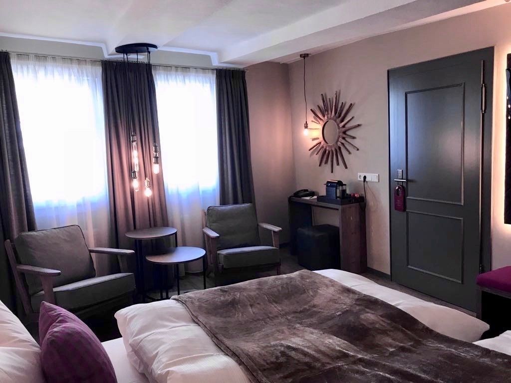 A hotel room with a bed, two armchairs, a small table, a desk, a mirror, and a door.