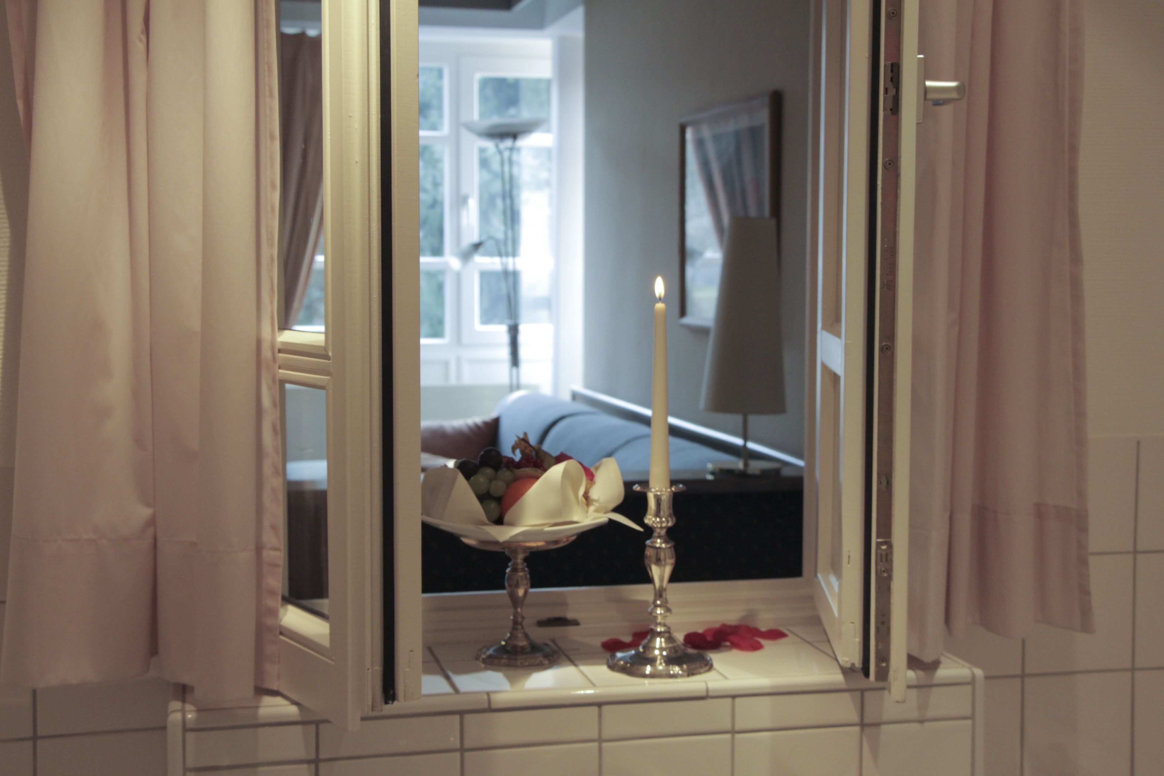 A bathroom with a mirror reflecting a room with a bed and a window.