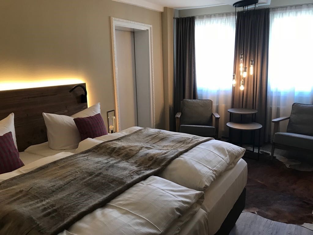 Een hotelkamer met een groot bed, een stoel, een tafeltje en uitzicht op het omliggende landschap, zichtbaar door gordijnen.