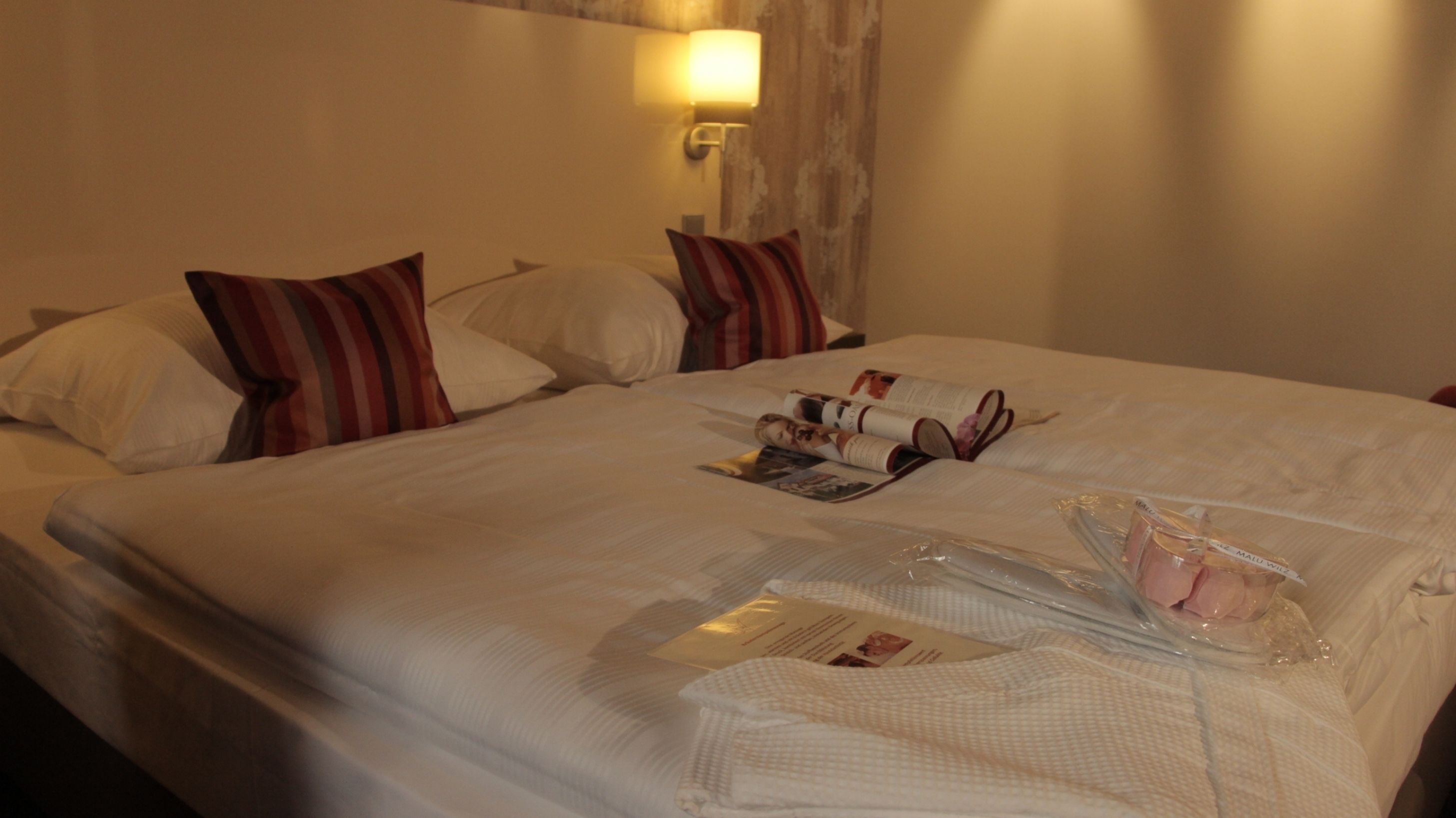 A hotel room with two beds, each with white bedding and striped pillows. There is a wall-mounted lamp and a small table with a magazine and a tissue box.