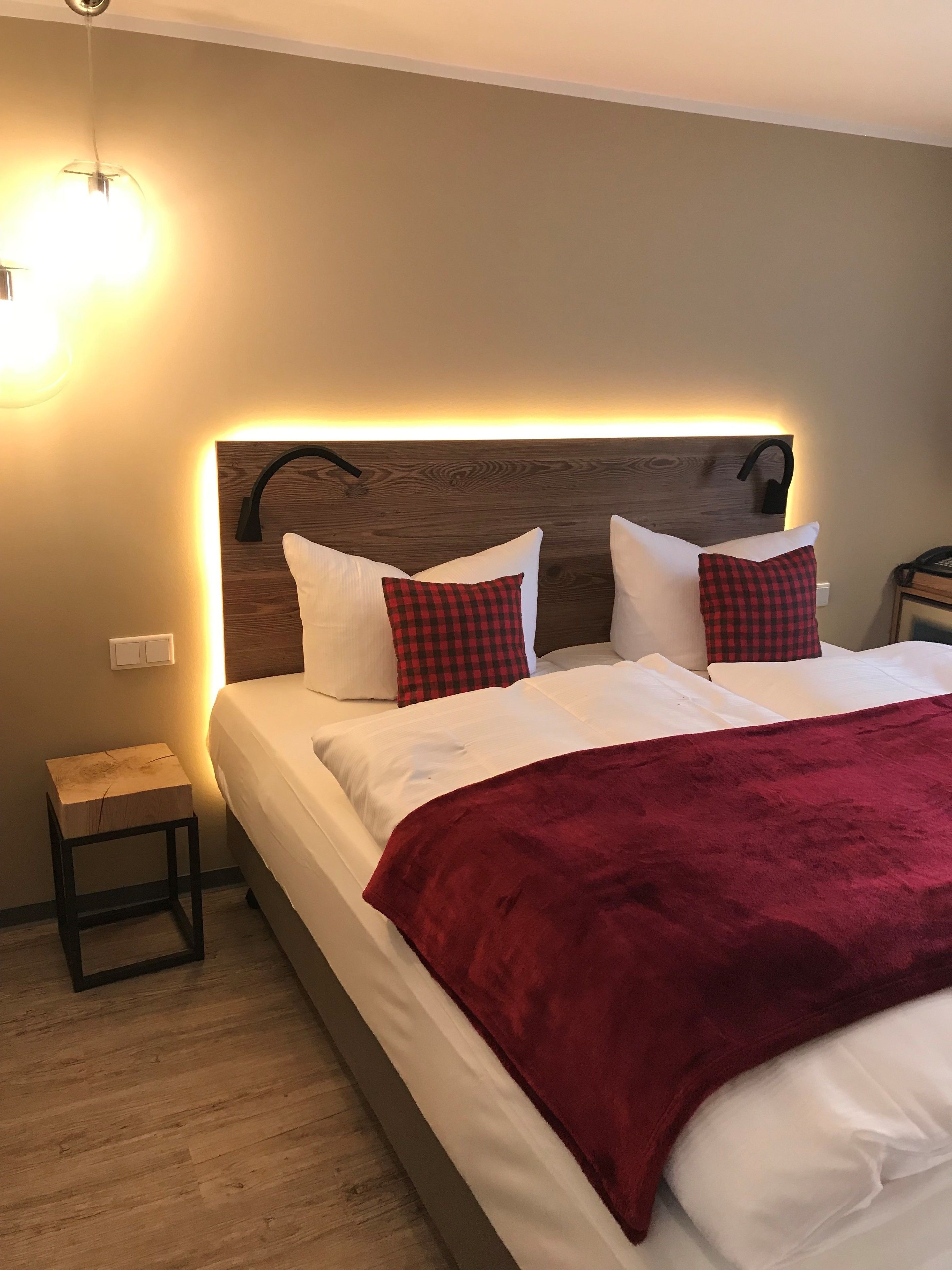 Ein ordentlich bezogenes Bett mit roter Decke, zwei karierten Kissen und einem Holzkopfstück mit integrierter Beleuchtung.