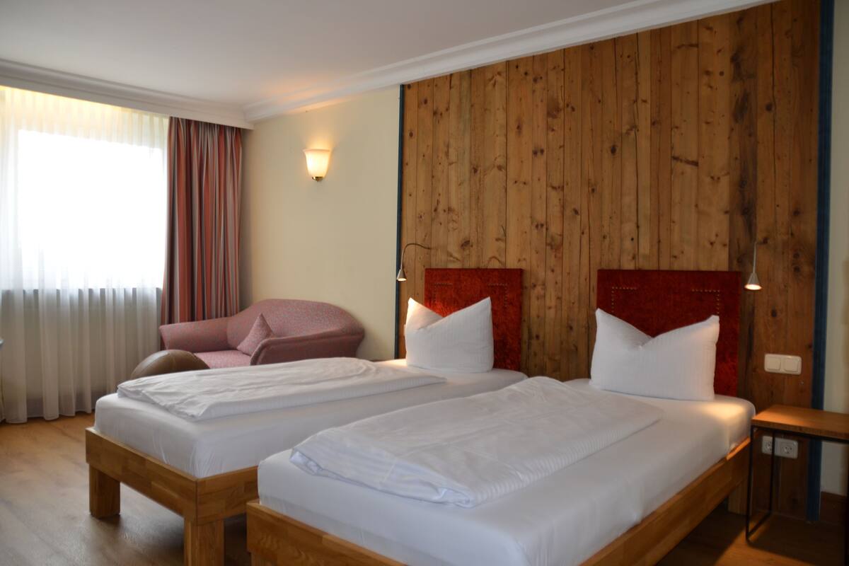 Een hotelkamer met twee enkele bedden, een houten hoofdeinde, een rode accentmuur, een houten bankje en een raam met gordijnen.