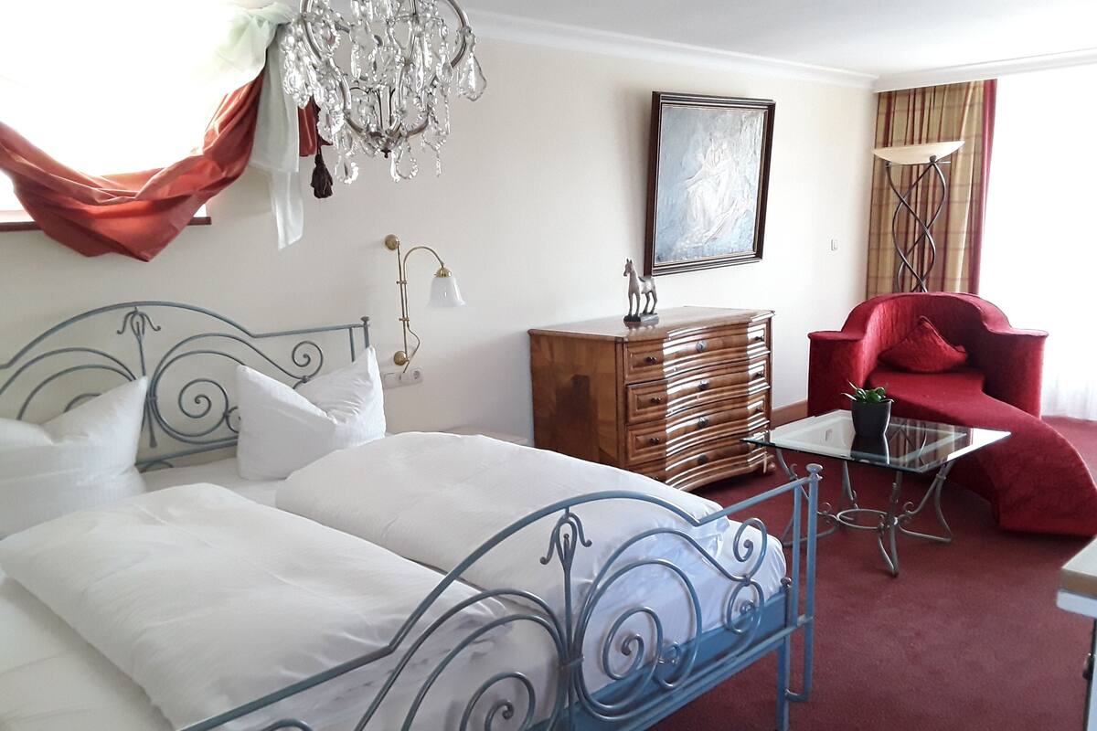 Een slaapkamer met een bed met metalen frame, een houten dressoir, een rode fauteuil, een glazen salontafel en een kroonluchter.
