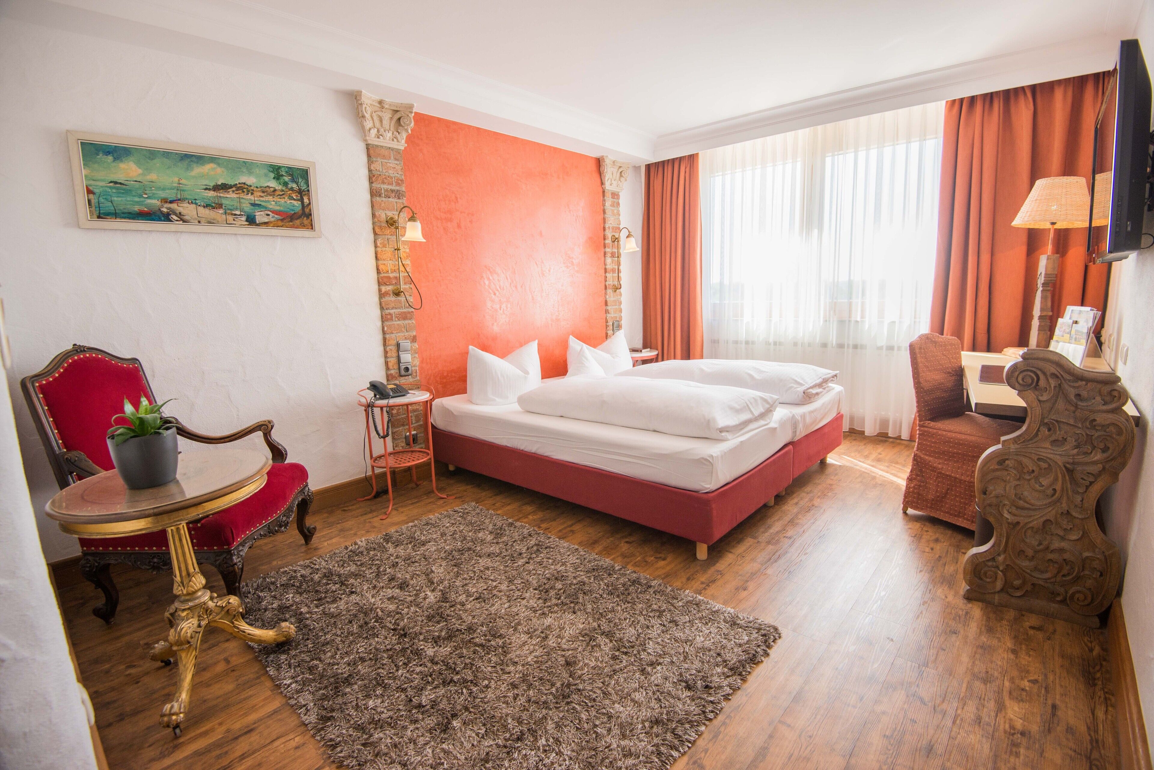 Habitación de hotel con una cama grande, una pared de acento rojo, un cuadro, un sillón rojo, una mesita con una planta y suelo de madera.