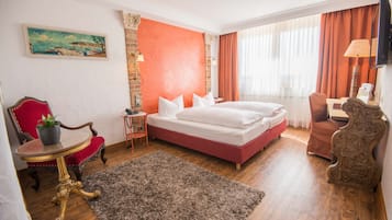 Habitación de hotel con una cama grande, una pared de acento rojo, un cuadro, un sillón rojo, una mesita con una planta y suelo de madera.