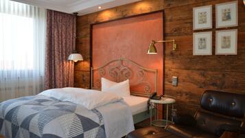 Quarto de hotel com cama, poltrona de couro, mesa de cabeceira com abajur e obras de arte na parede.