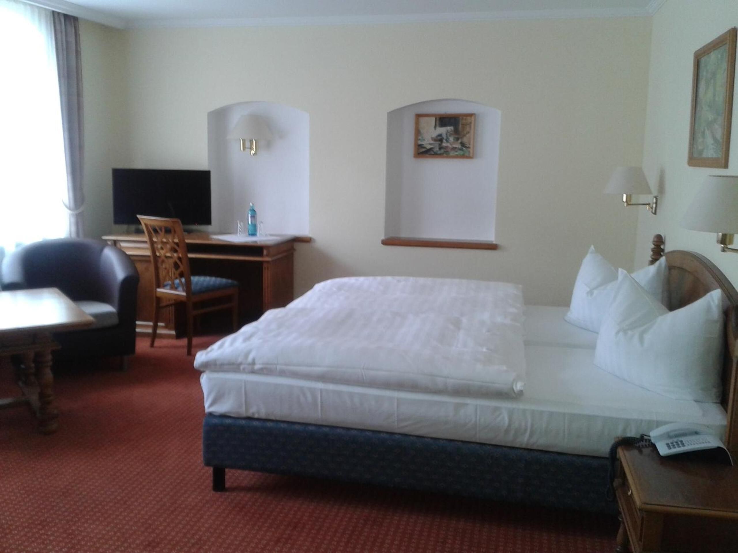 A hotel room with a bed, a desk with a chair, a television, and a painting on the wall.