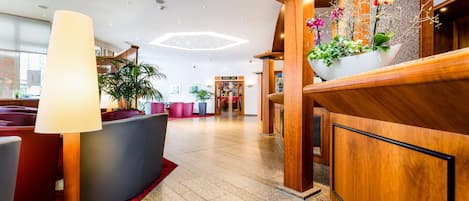 A modern lobby with a reception desk, a large floor lamp, a potted plant with flowers, and a seating area with red and grey chairs.