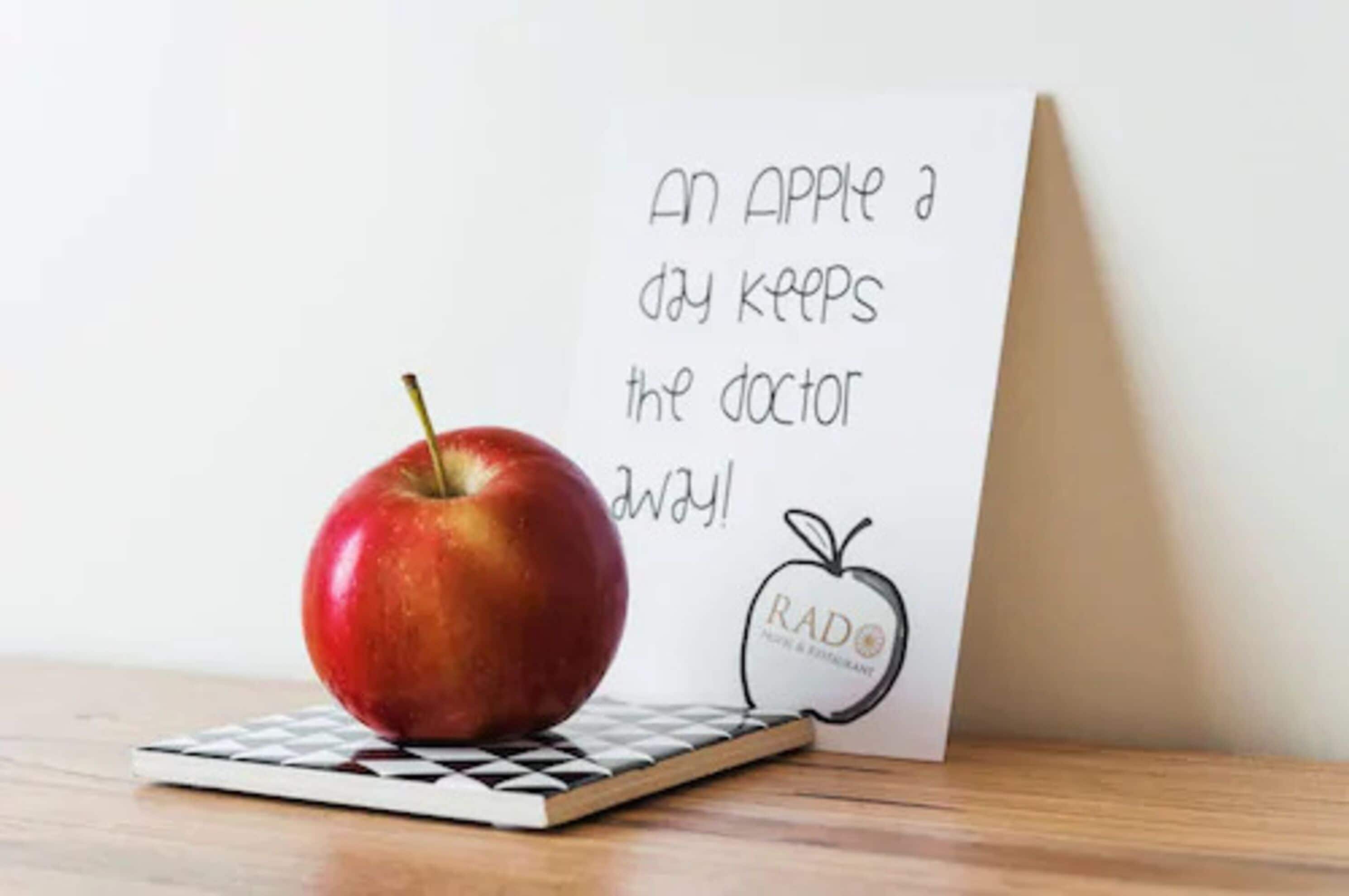 Una manzana roja sobre un posavasos a cuadros junto a una tarjeta con un dibujo de manzana y texto.