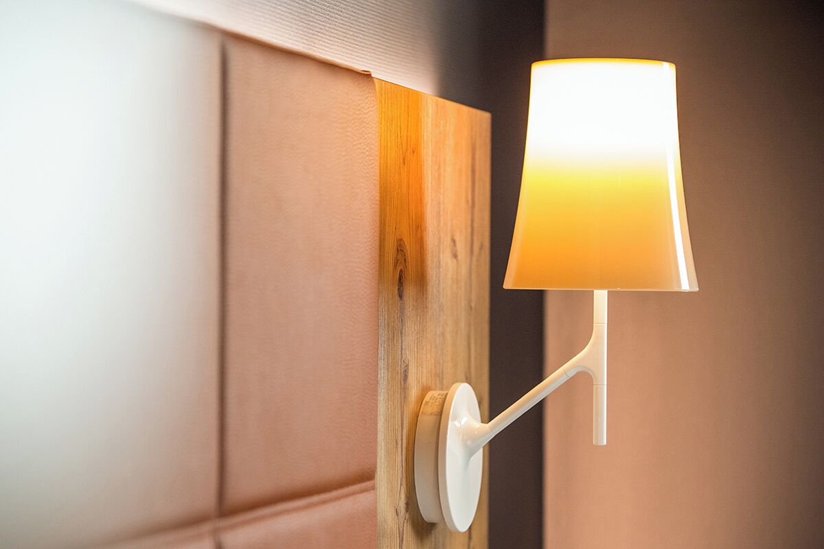 Una lámpara de pared con base blanca y pantalla cilíndrica, fijada a un panel de madera sobre un fondo de pared beige.