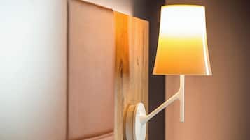 Una lámpara de pared con base blanca y pantalla cilíndrica, fijada a un panel de madera sobre un fondo de pared beige.