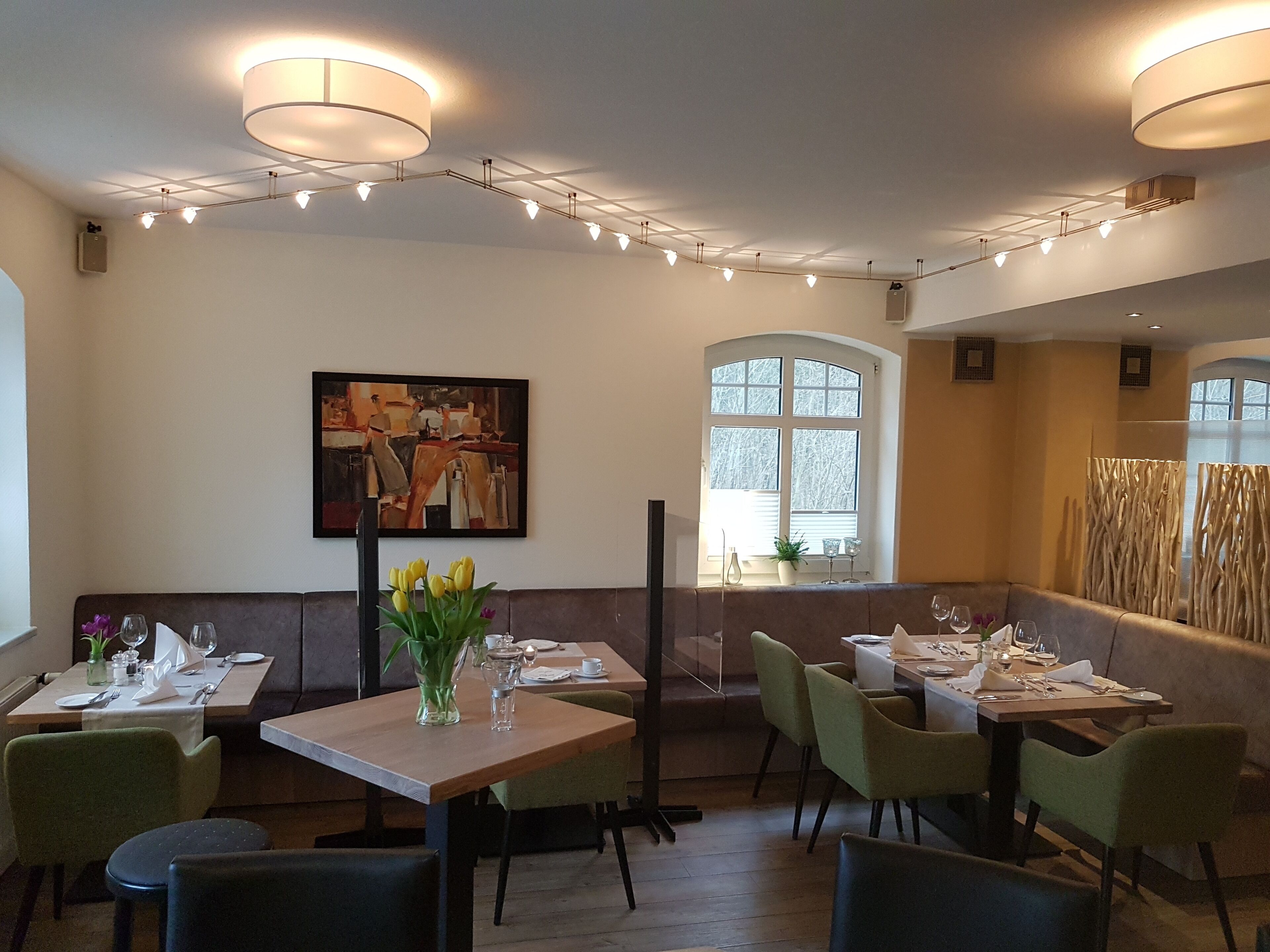 A restaurant interior with tables set for dining, a painting on the wall, and a window with a view.