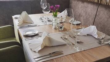 Een eettafel gedekt voor twee personen, met een vaas paarse bloemen, heldere glazen en netjes gevouwen servetten.