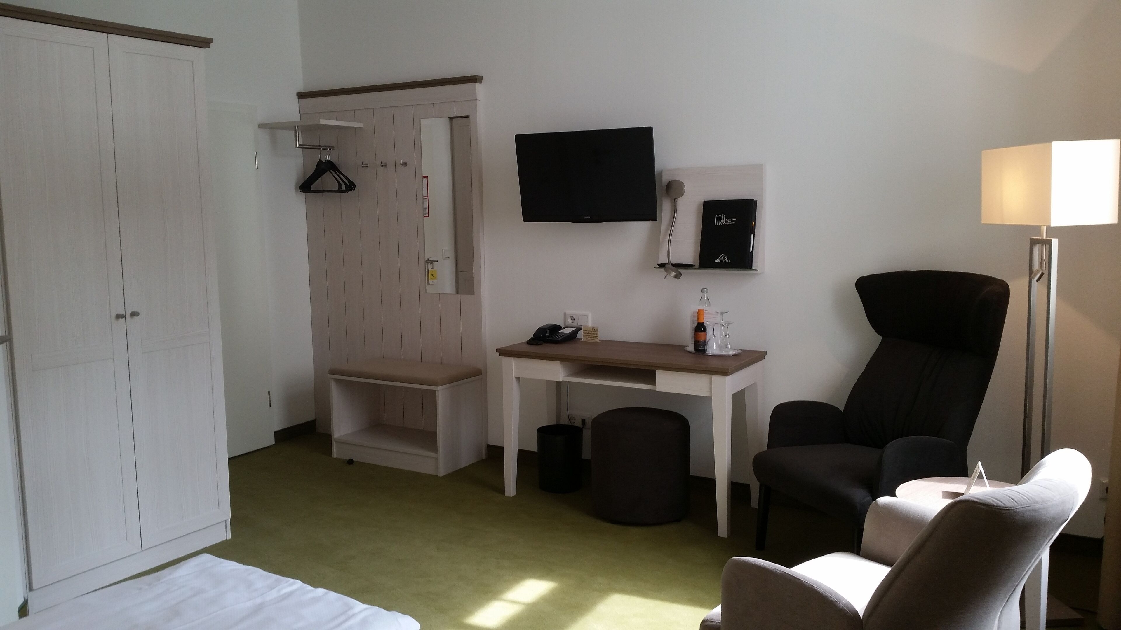 Ein Hotelzimmer mit einem Bett, einem Schreibtisch, einem Sessel, einem Kleiderschrank, einem Fernseher, einer Lampe und einer Weinflasche.
