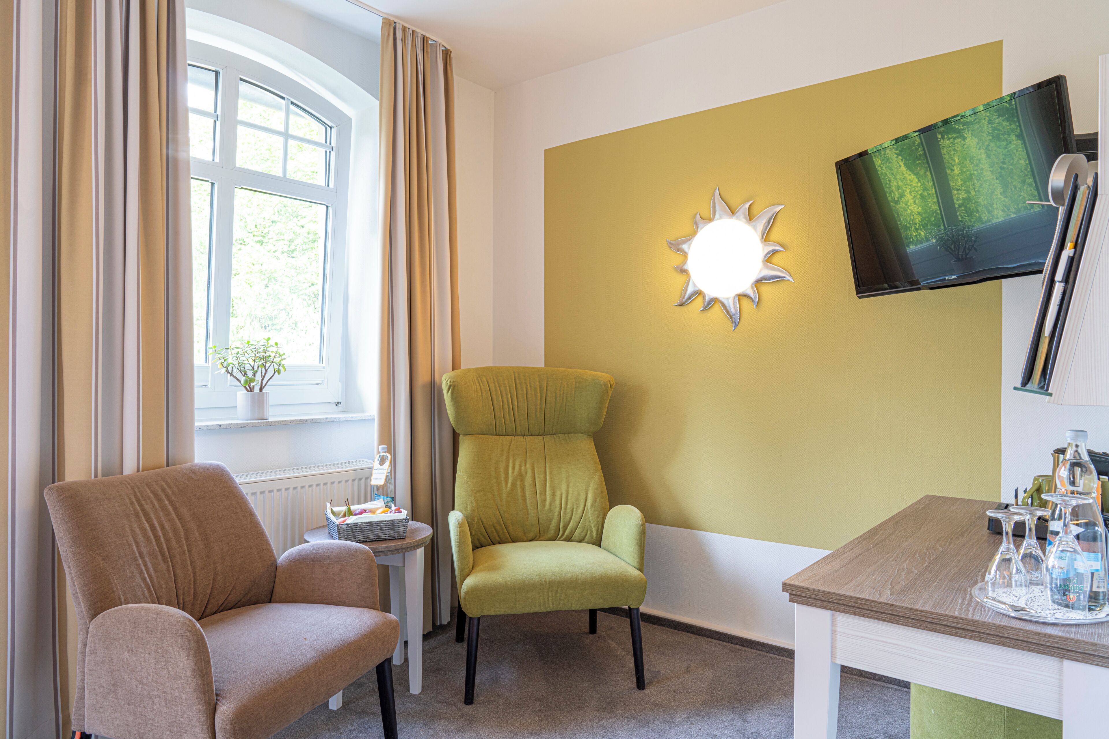 Une pièce comprenant un canapé, un fauteuil, une petite table avec un plateau, un téléviseur fixé au mur et une fenêtre avec des rideaux.