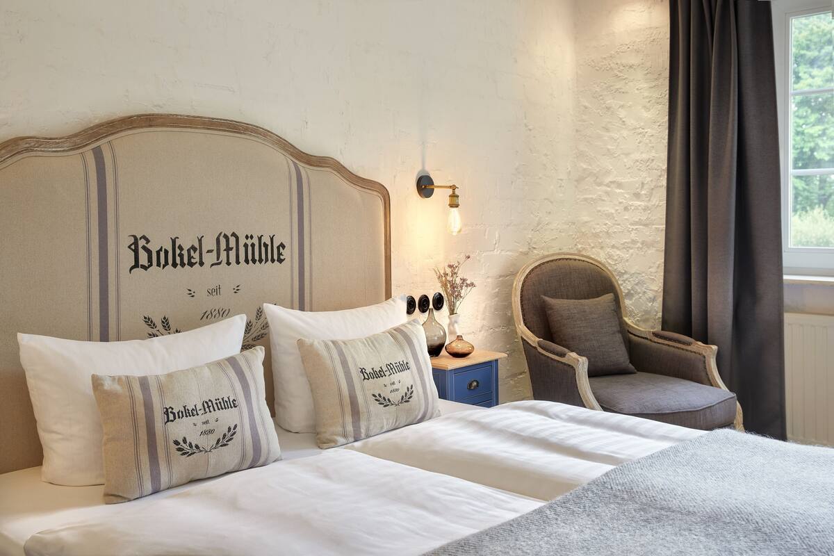 Una cama bien hecha con dos almohadas, una mesita de noche con una lámpara y una silla en una habitación con pared texturizada y una ventana con cortinas.