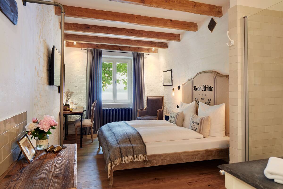 Un dormitorio con una cama de madera, una mesita de noche con un jarrón de flores, una silla y una ventana con cortinas.