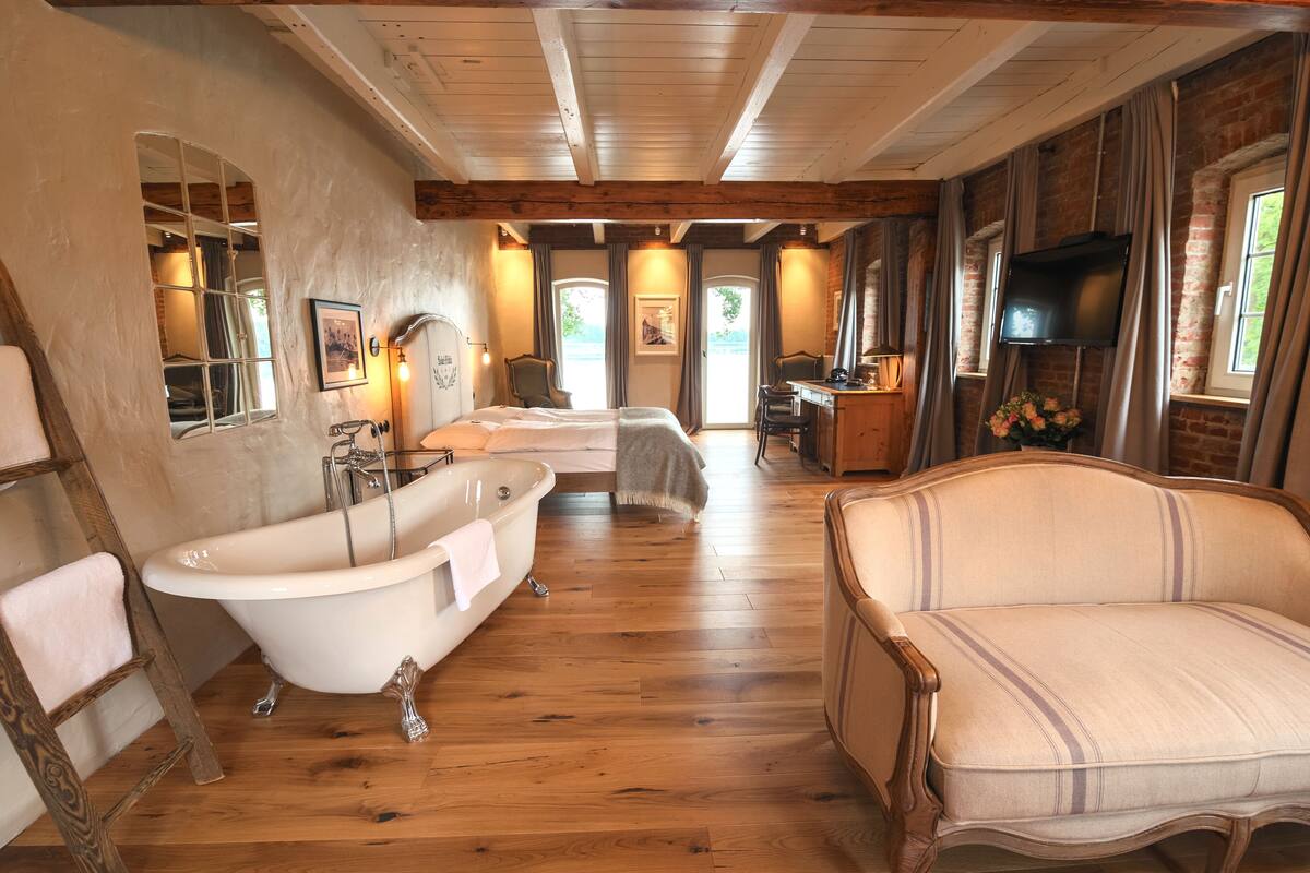 Una habitación con piso de madera, una bañera, un sofá, un televisor y una ventana con cortinas.
