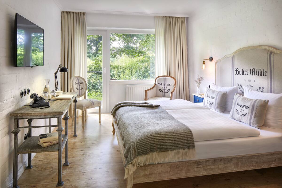 Un dormitorio con una cama grande, un escritorio y una silla. Hay una ventana con cortinas, un radiador y un televisor montado en la pared.