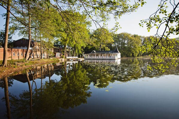 A riverside building with a dock, surrounded by trees and a calm water body. - Bokel-Mühle am See (Bokel)
