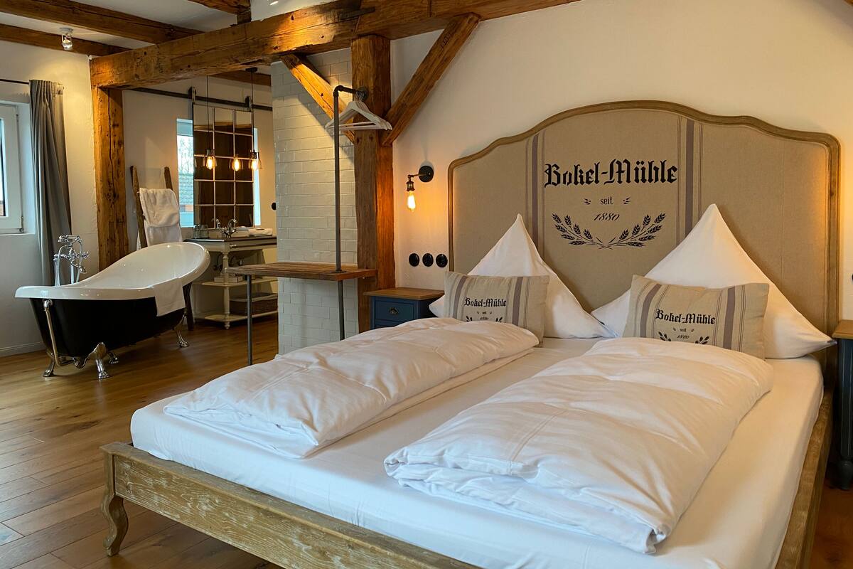 Habitación de hotel con una cama grande, un banco de madera, una bañera y un espejo.