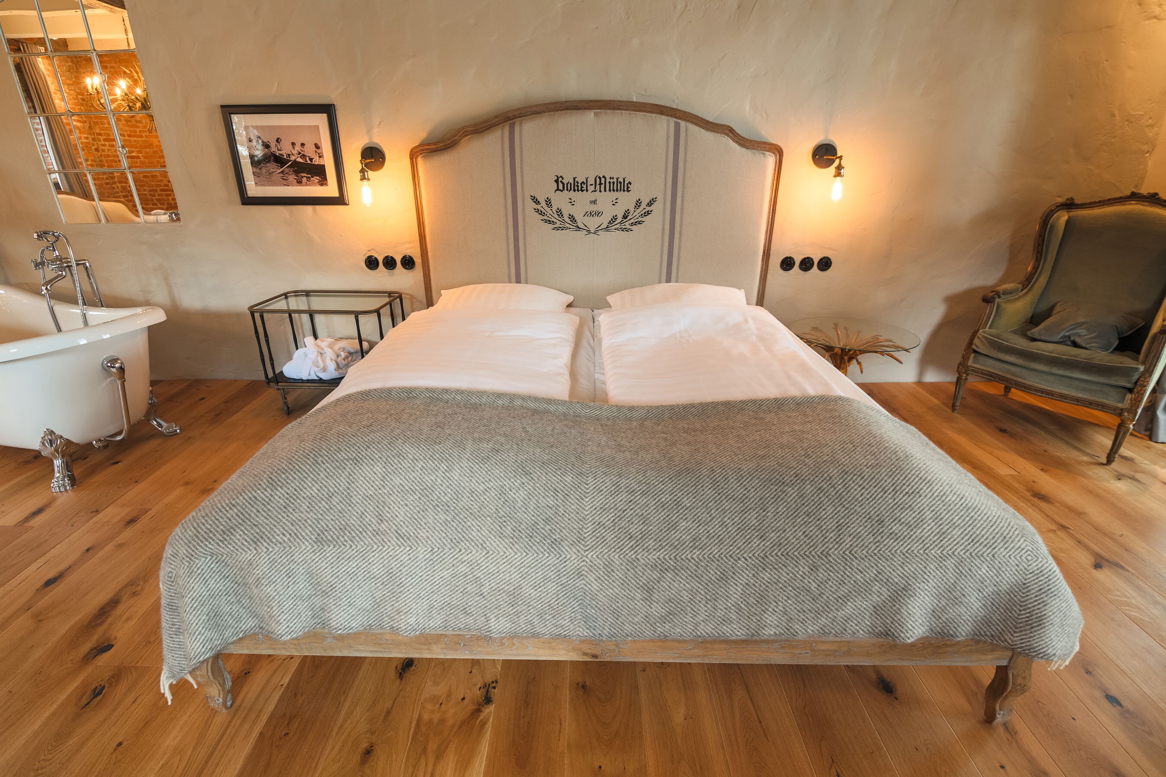 Une chambre avec un grand lit, un plancher en bois, une tête de lit de style vintage, un tableau accroché au mur et une chaise.