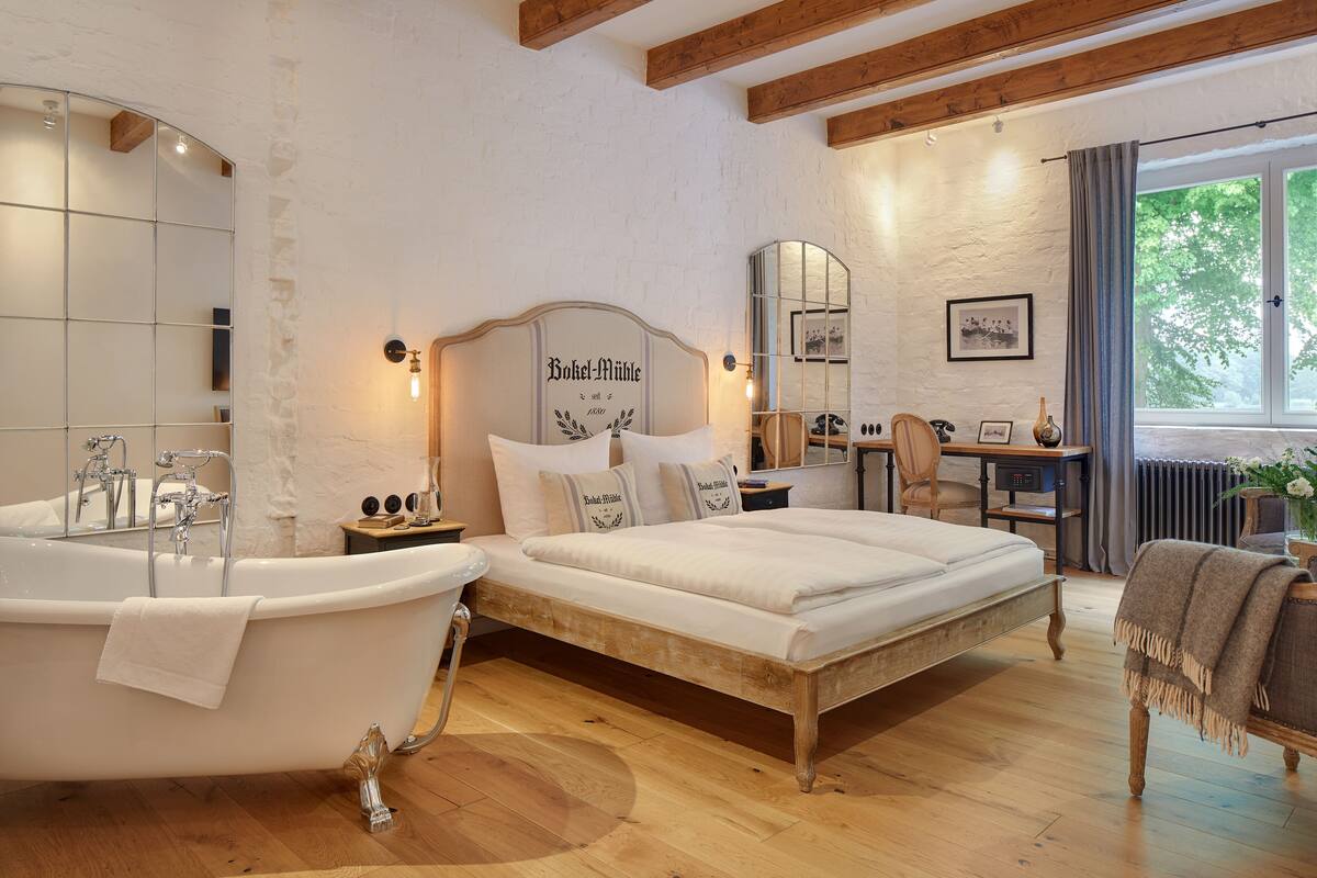 Un dormitorio con una cama grande, una bañera, pisos de madera, un escritorio y una ventana con vista a árboles.