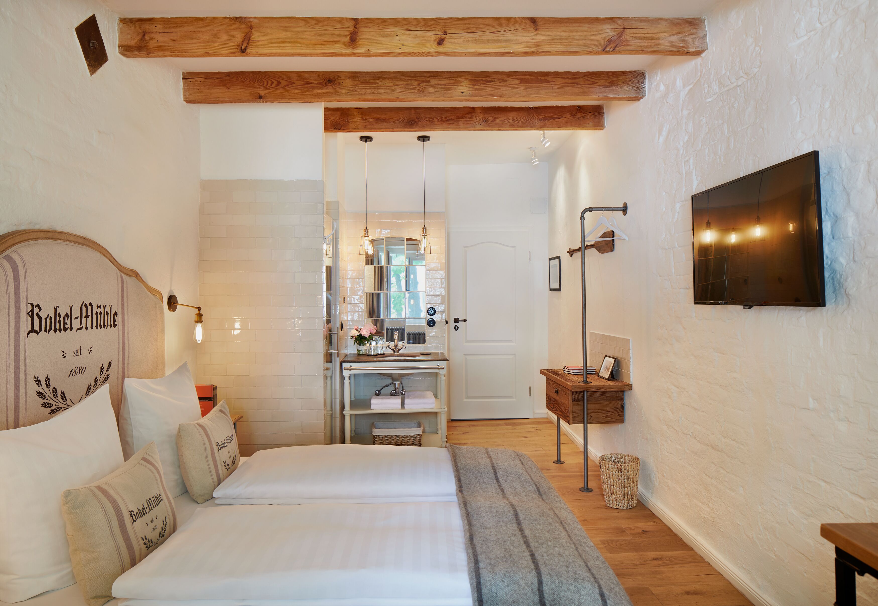 Un dormitorio con una cama doble, cabecera de madera, mesita de noche, televisor montado en la pared y una cocina visible a través de una puerta abierta.