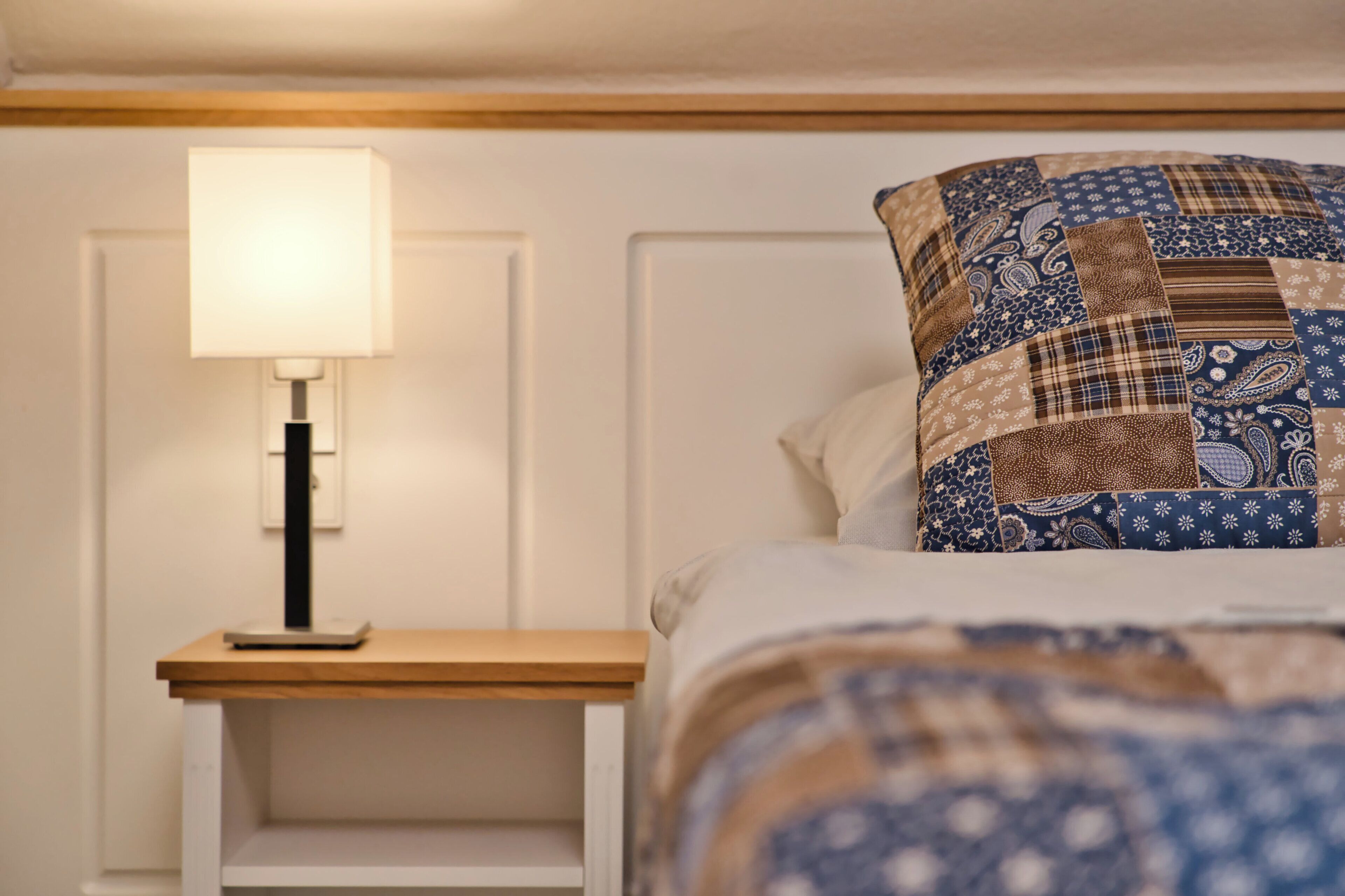 Una cama bien hecha con un edredón estampado, una lámpara de noche y una mesita de noche blanca.