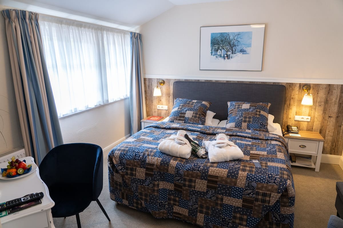 Habitación de hotel con una cama grande, dos mesitas de noche, una silla, una pequeña mesa con fruta y controles remotos, y un cuadro en la pared.