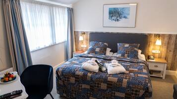 Habitación de hotel con una cama grande, dos mesitas de noche, una silla, una pequeña mesa con fruta y controles remotos, y un cuadro en la pared.