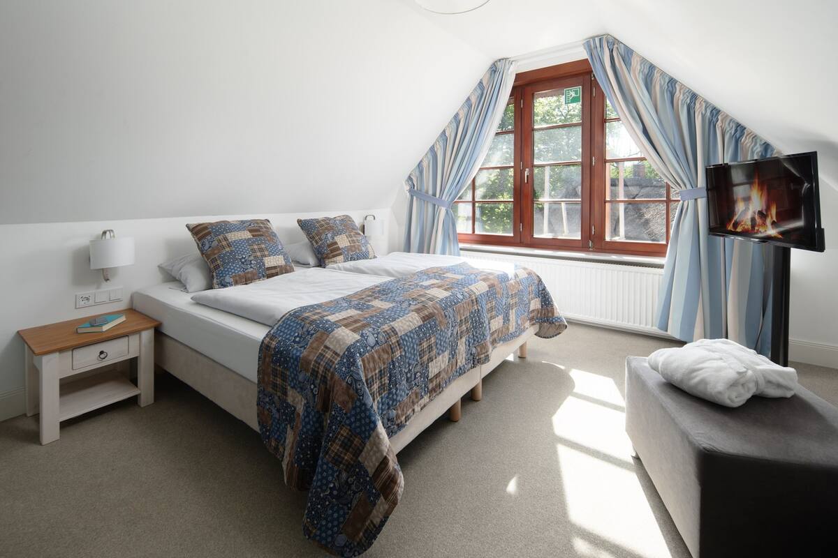 Un dormitorio con cama, mesita de noche, lámpara, ventana con cortinas y televisor.