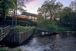Ein Fluss mit einer Brücke, ein Gebäude mit großen Fenstern und eine von Bäumen gesäumte Uferzone.