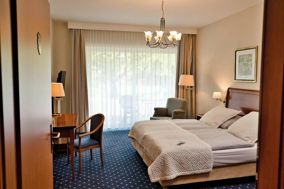 Una habitación de hotel con una cama, dos sillas, un escritorio, una araña de cristal y una ventana con cortinas.