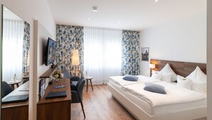 Una habitación de hotel con cama, escritorio con una computadora portátil, una silla, televisor y ventana con cortinas.
