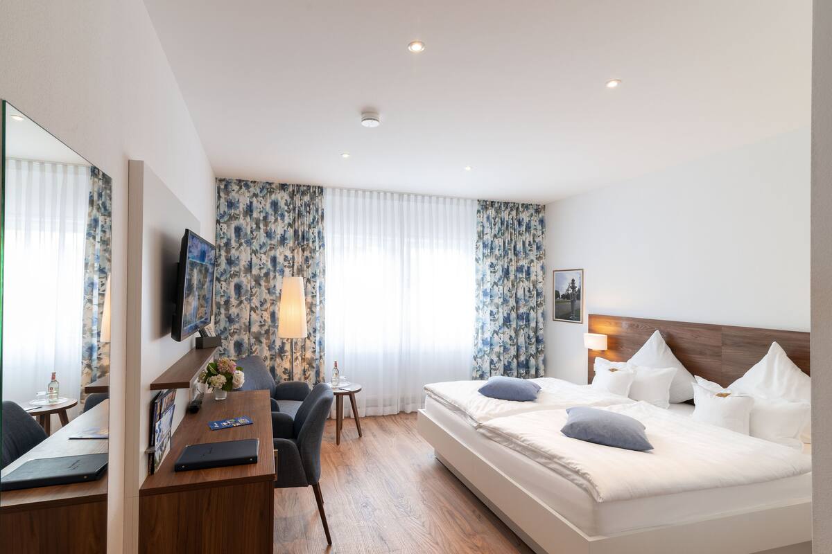 A hotel room with a bed, a desk with a laptop, a chair, a TV, and a window with curtains.