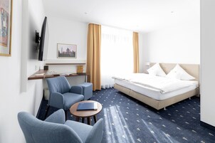 A hotel room with a bed, two armchairs, a desk, a TV mounted on the wall, and a window with curtains.
