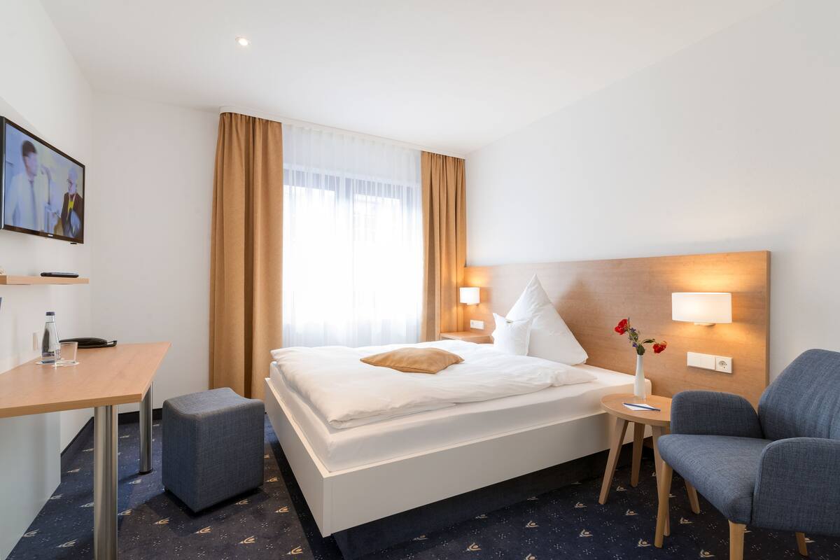 A hotel room with a bed, bedside table, chair, and a TV mounted on the wall.