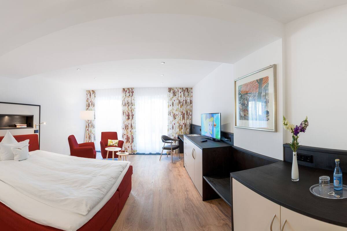 A hotel room with a bed, a desk, a TV, and a small table with a vase of flowers.