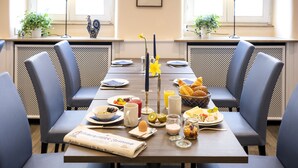 Un comedor con una mesa preparada para cuatro personas, con un periódico, alimentos para el desayuno y un jarrón con flores.