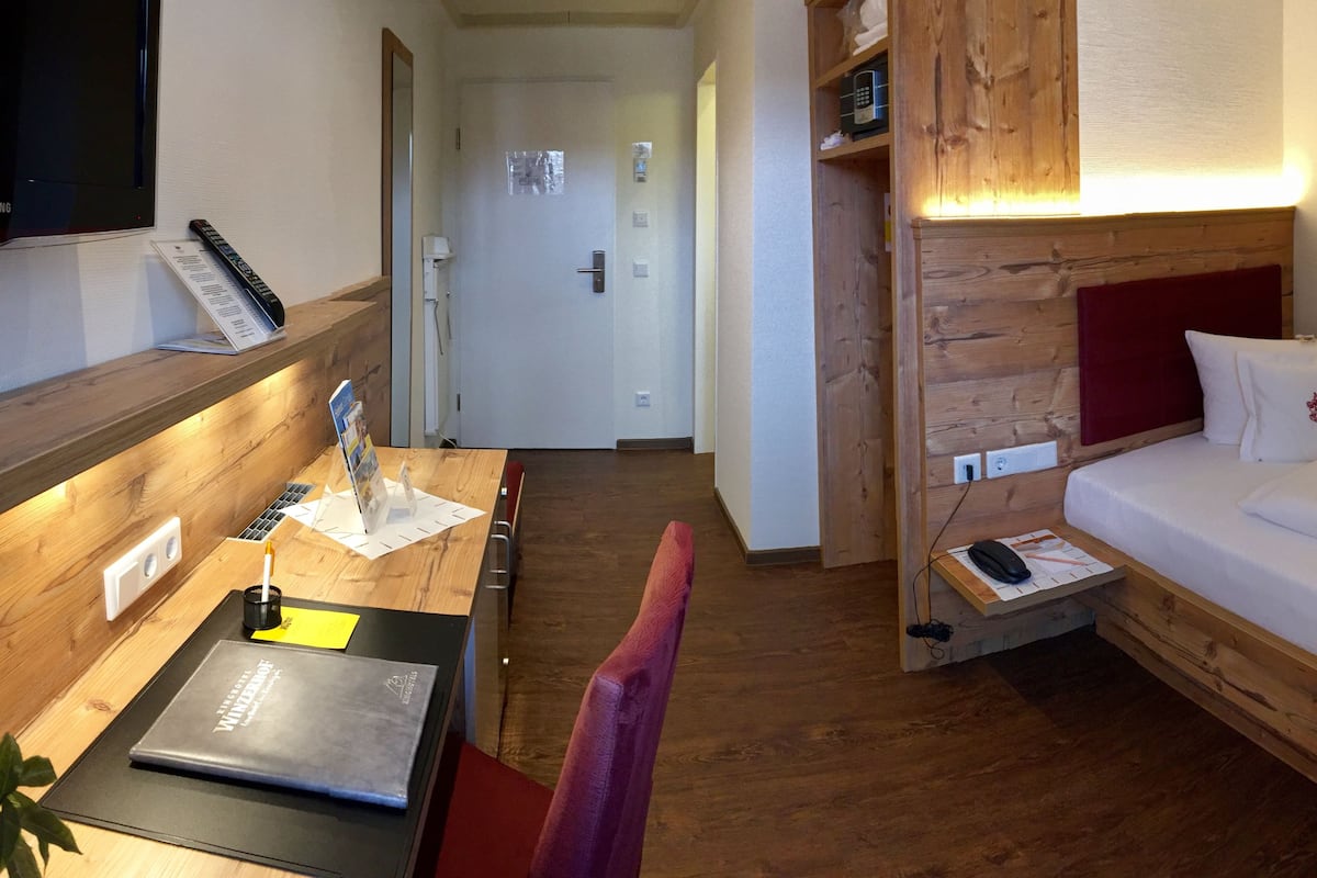 Habitación de hotel con cabecera de madera, cama con ropa blanca, escritorio con una computadora portátil y un libro, una silla y una mesita con un control remoto y un menú.