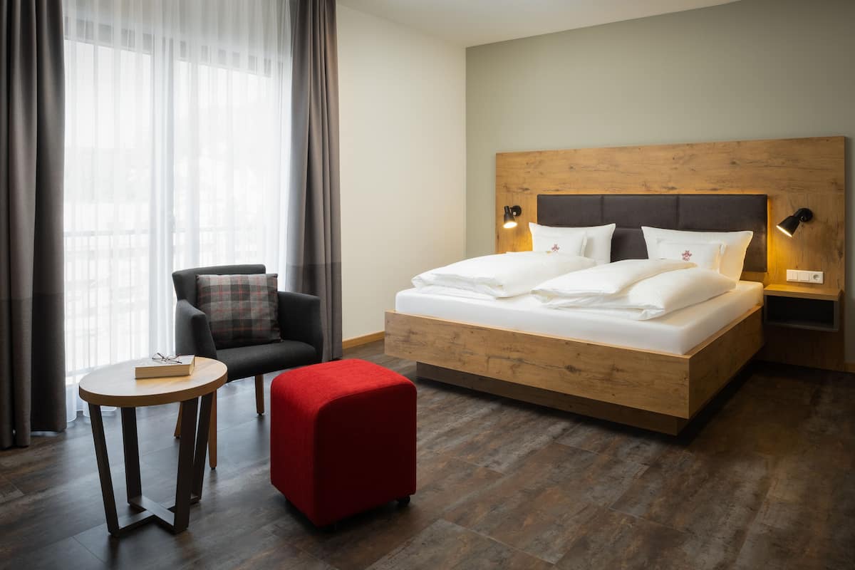 Habitación de hotel con cabecera de madera, una cama individual, un reposapiés rojo, una mesita redonda pequeña, una silla y una mesita de noche con lámparas.