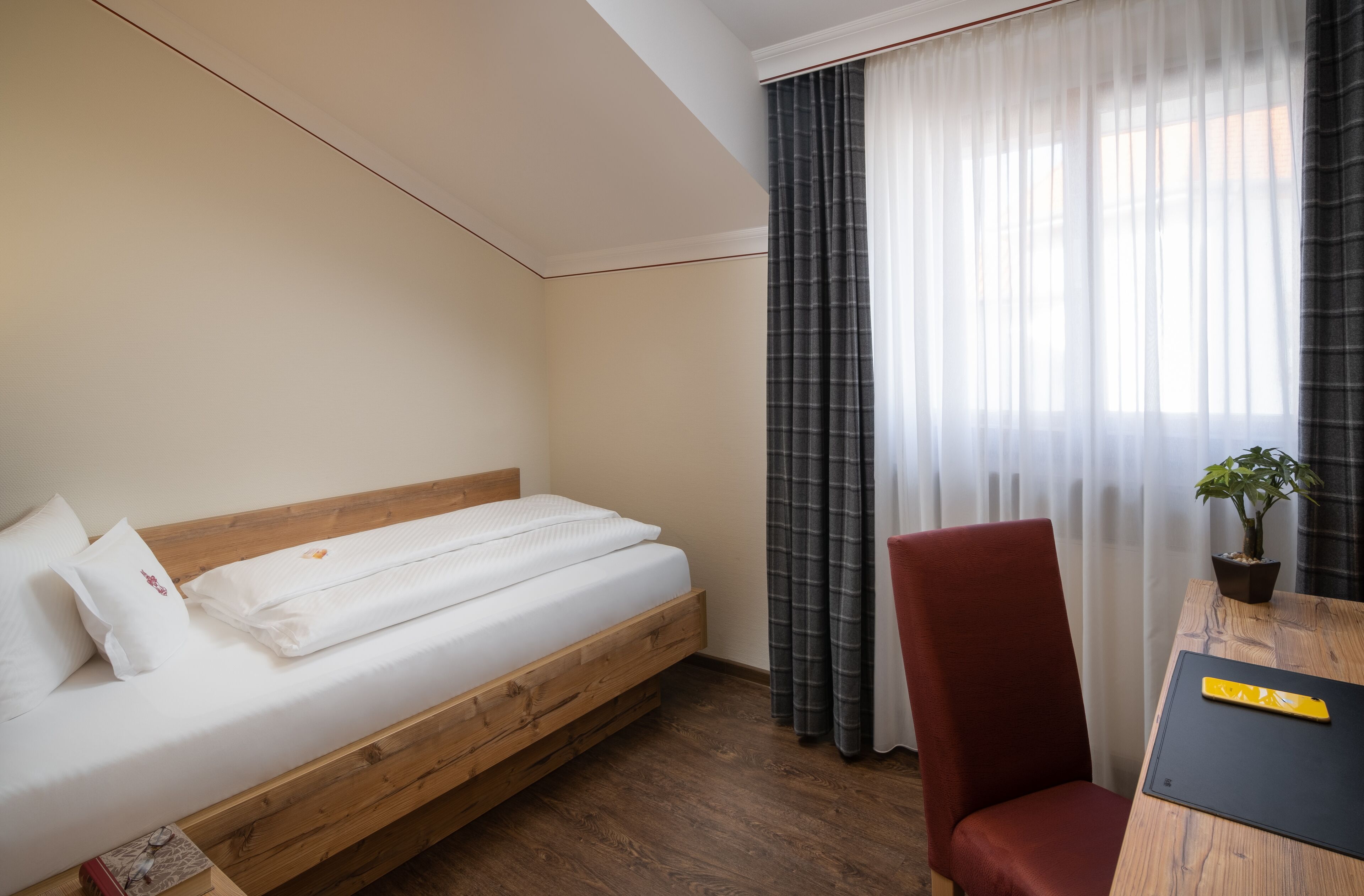 Habitación de hotel con una cama de madera, una silla roja, una mesita con una planta y cortinas traslúcidas.